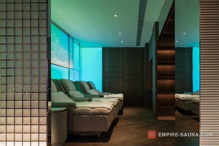 Row of pale recliner chairs in the lounge at Empire Sauna, Macau, facing a turquoise backlit wall beside a glass-brick screen