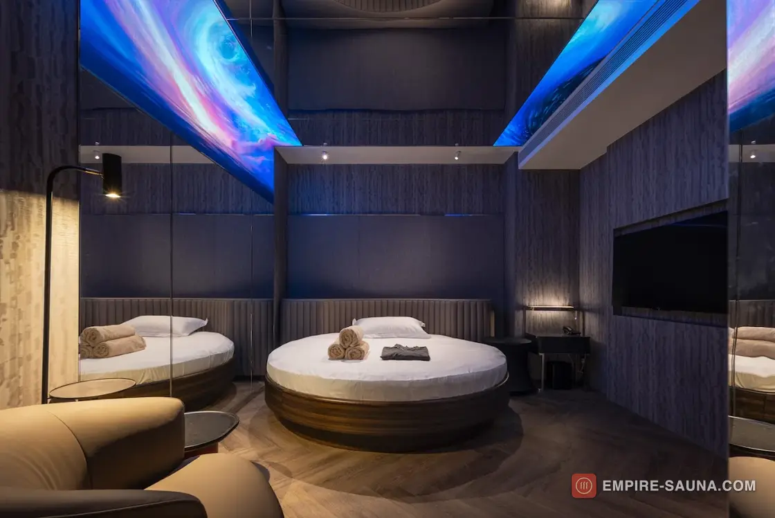 Round bed on a dark timber plinth in a themed suite at Empire Sauna, Macau, under a blue and violet lit ceiling panel, with a herringbone floor and a mirrored wall